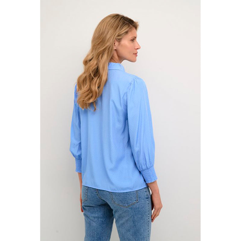 Blå gennemknappet bluse med struktur i stoffet den har almindelig skjortekrave og lange ærmer som afsluttes med smock set bagfra
